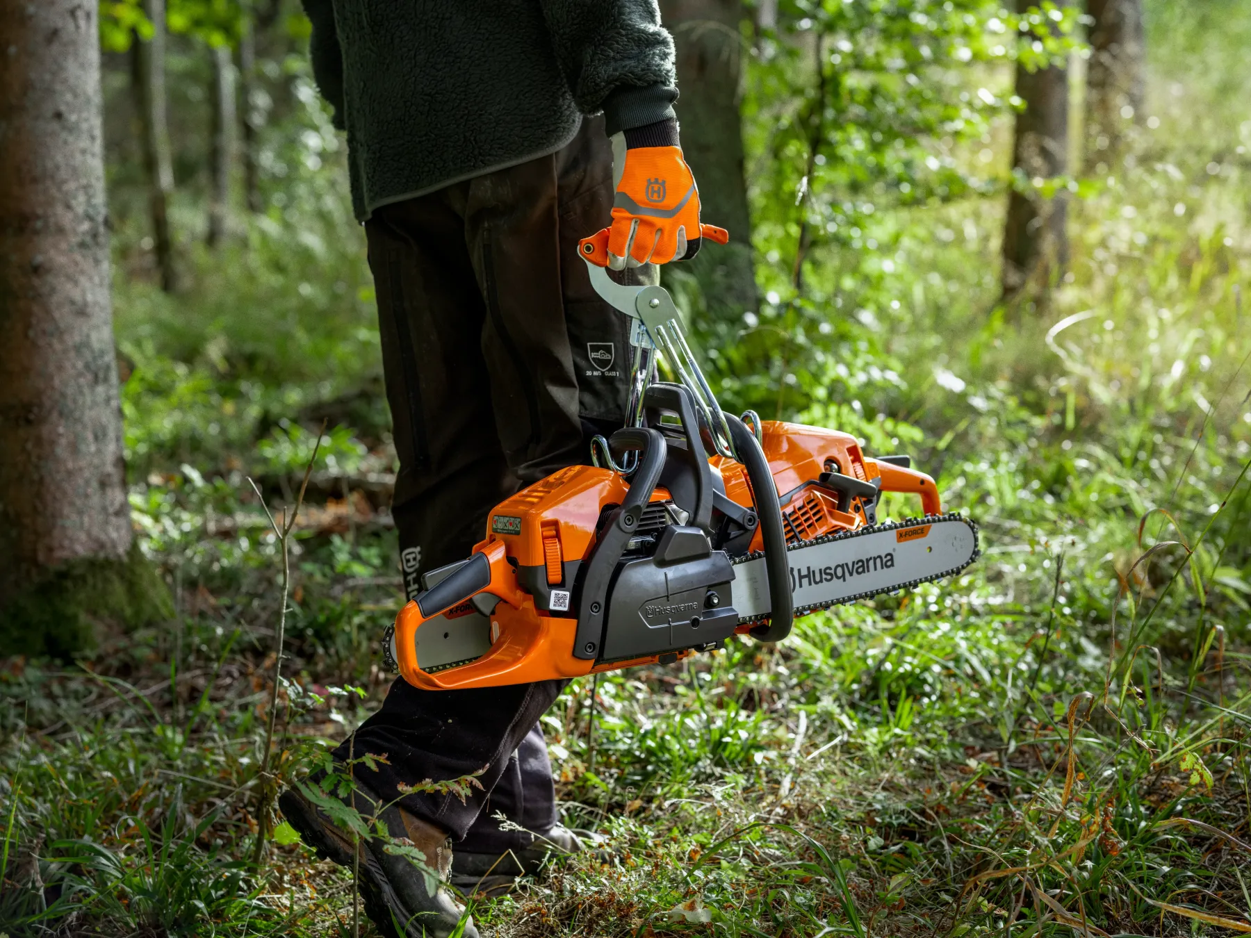 Motorsåg En person iförd handskar och friluftskläder bär två orange Husqvarna-motorsågar i en grön skog och går genom gräs och träd.