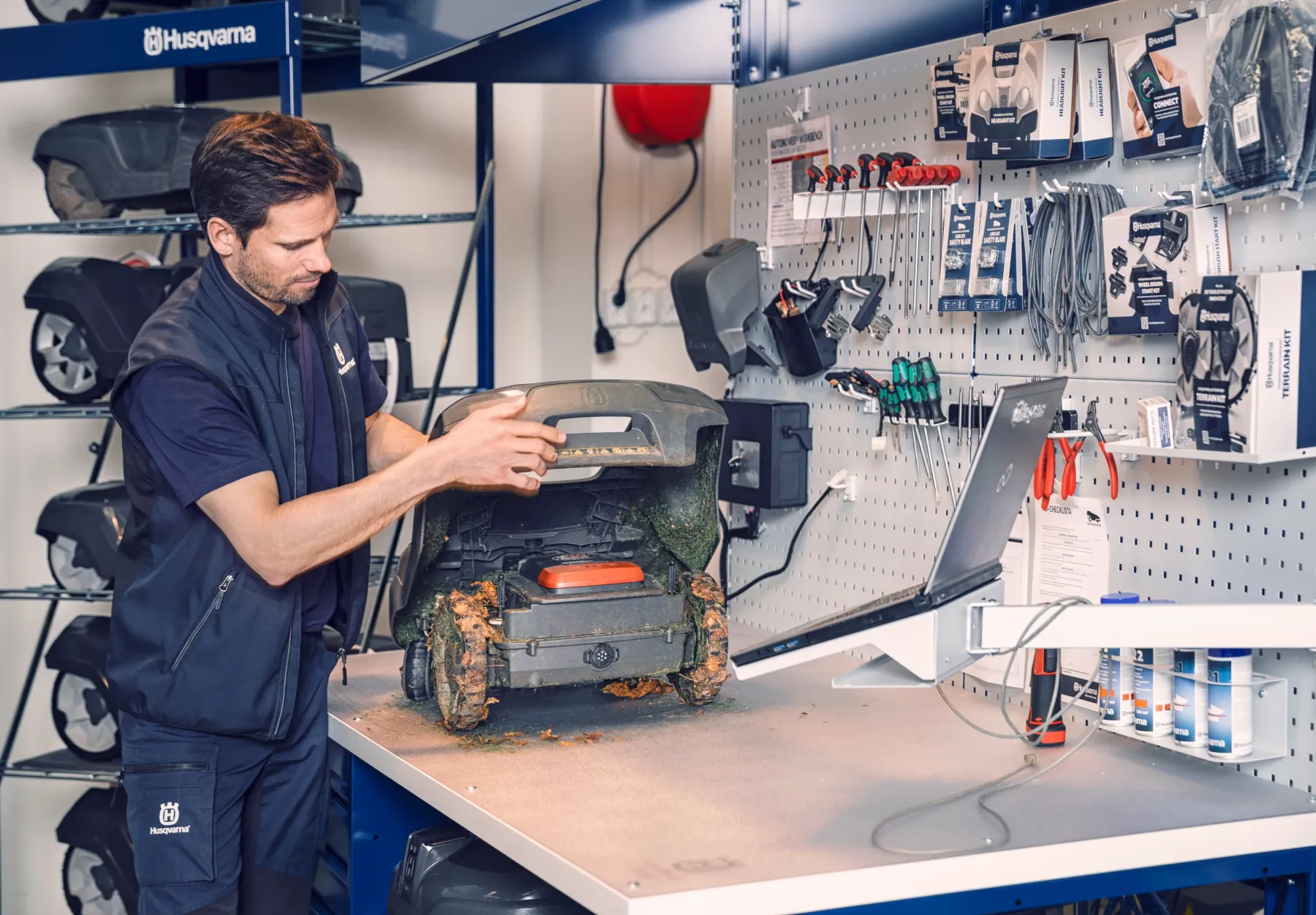 H750-0052. En tekniker i Husqvarnauniform reparerar en robotgräsklippare vid en arbetsbänk i en välorganiserad verkstad med verktyg, kablar och reservdelar hängande på en pegboard.