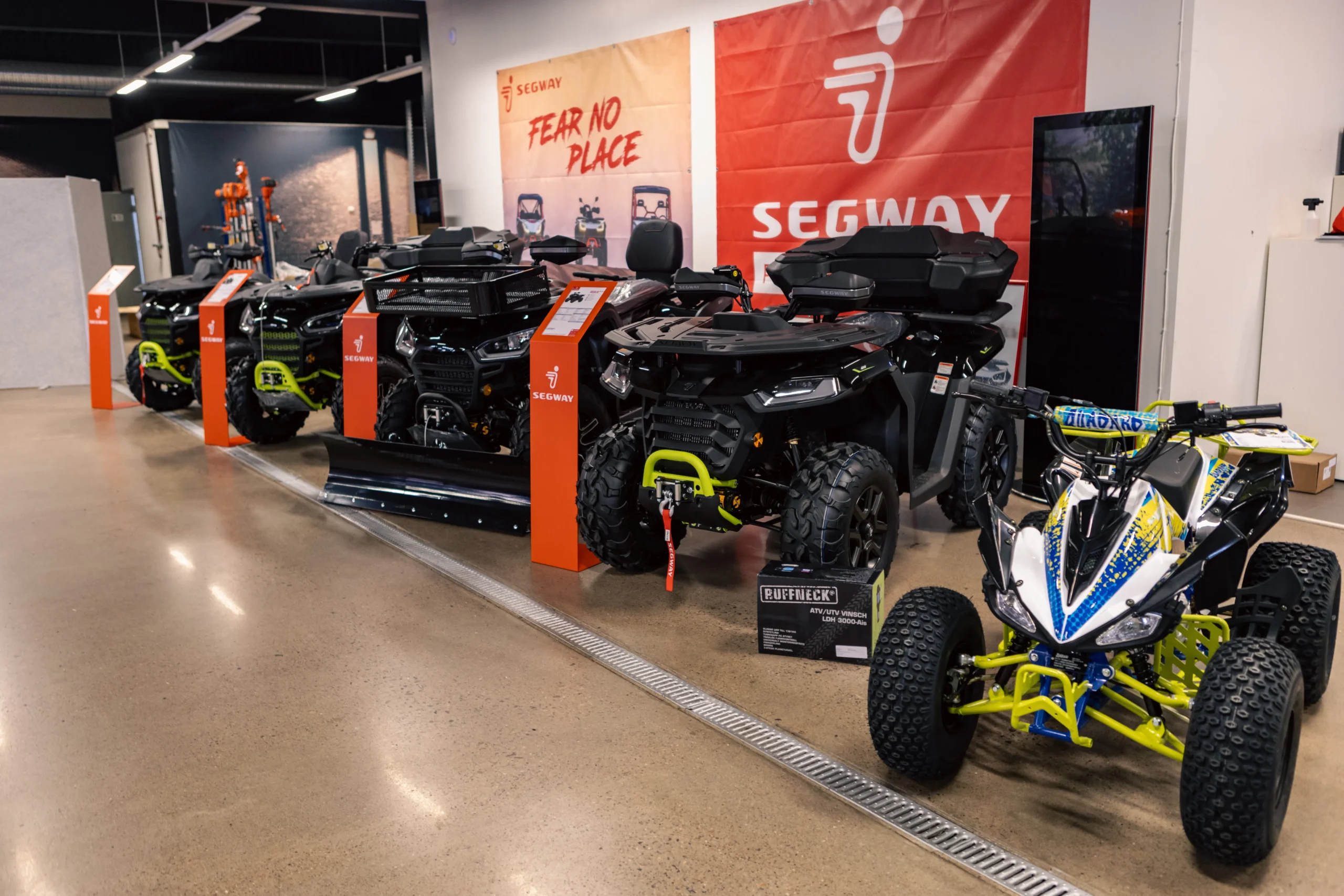 I en utställningslokal står flera svarta terrängfordon av märket Segway på rad, med orange Segway-skyltar och -banderoller bakom, och en mindre vit och blå fyrhjuling till höger.