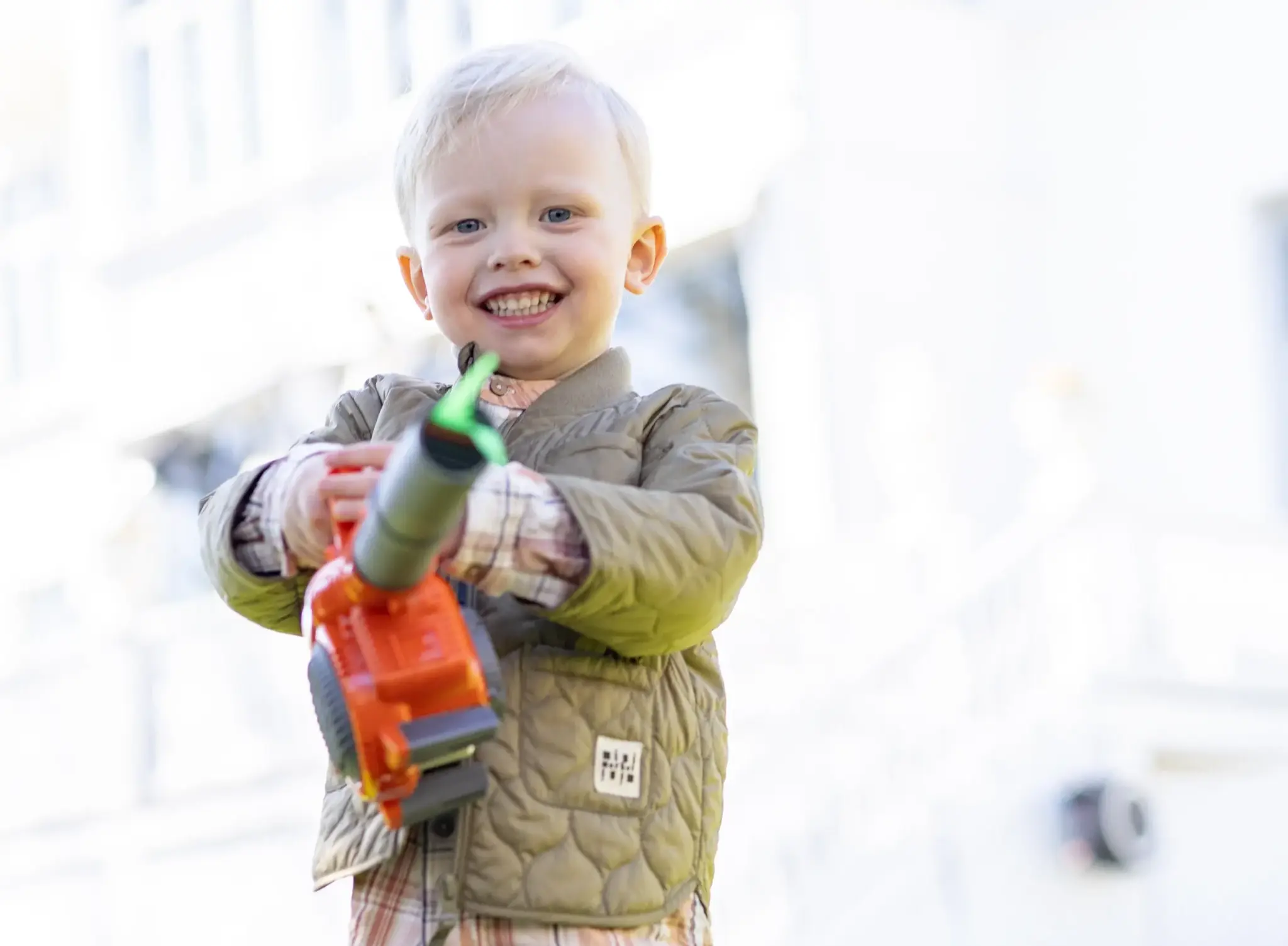Ett leende barn med blont hår håller i en stor lövblås utomhus, iklädd en quiltad jacka och rutig skjorta, med en suddig byggnad i bakgrunden.