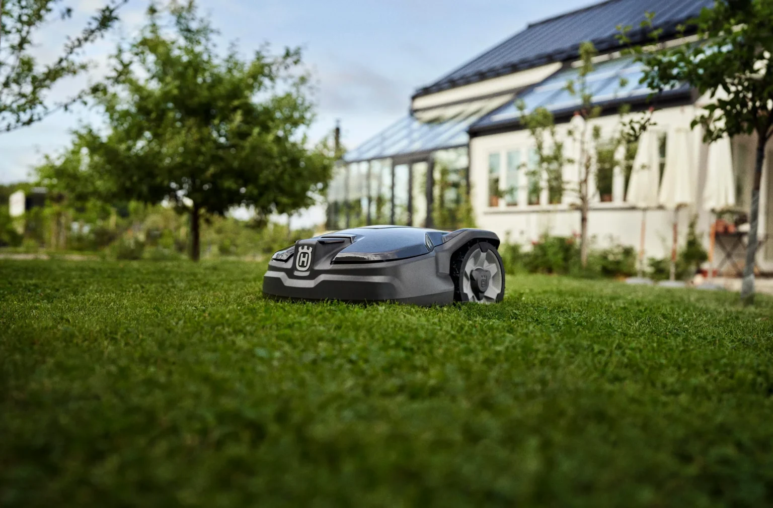 En robotgräsklippare klipper gräs i en trädgård nära ett hus med en växthustillbyggnad i glas. Träd och grönska omger området en solig dag.