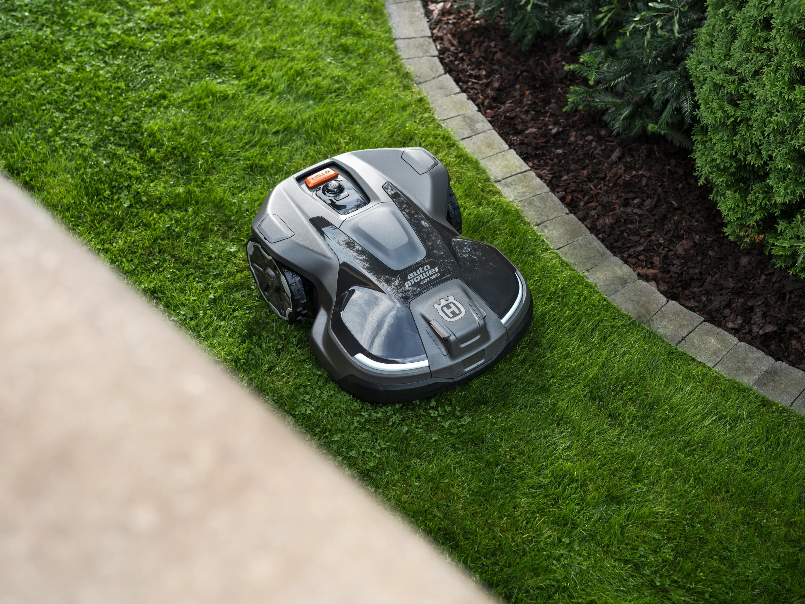 En robotgräsklippare arbetar på en grön gräsmatta nära en trädgårdsrabatt kantad med tegelstenar och buskar, sett uppifrån.