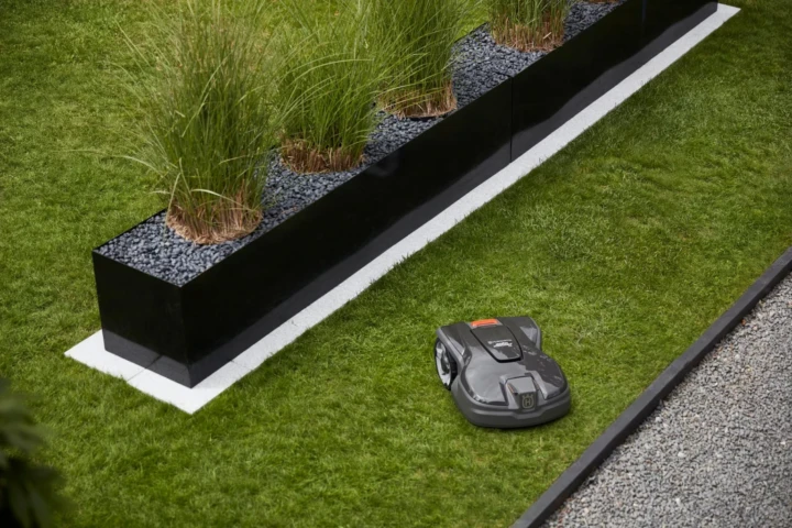 En robotgräsklippare trimmar gräset längs kanten av en modern trädgård med en upphöjd svart planteringskärl fylld med höga gröna växter och mörkt grus.