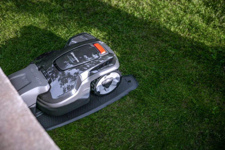 En robotgräsklippare är placerad på grönt gräs intill en sten- eller betongkant, delvis i skuggan, och förbereder sig för att klippa gräsmattan.
