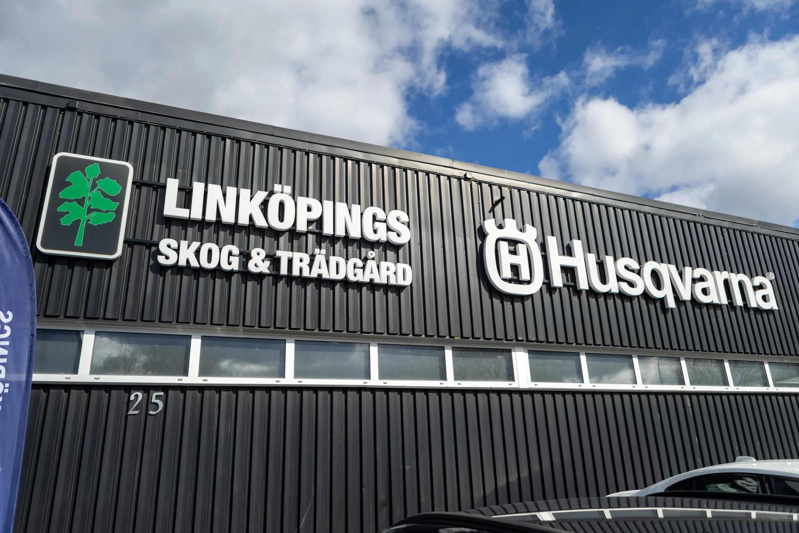 Connys Skog & Trädgård En svart byggnadsfasad med skyltarna "LINKÖPINGS SKOG & TRÄDGÅRD" och "Husqvarna" i vita bokstäver, tillsammans med deras logotyper. Himlen med moln syns ovanför byggnaden.