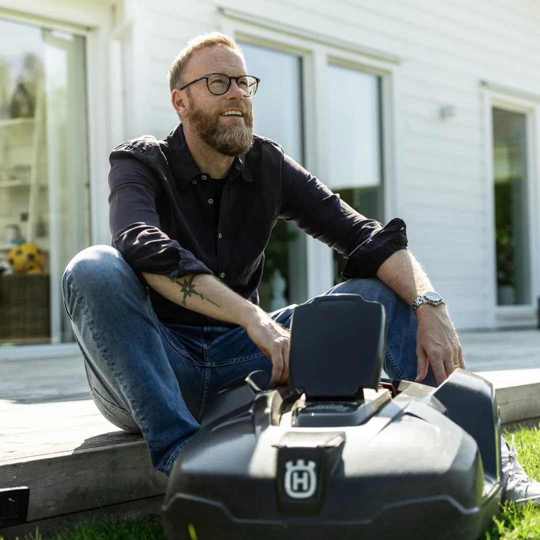 En leende man med glasögon och skägg sitter på ett trädäck bredvid sin personliga robotgräsklippare, med ett vitt hus och en grön gräsmatta i bakgrunden.