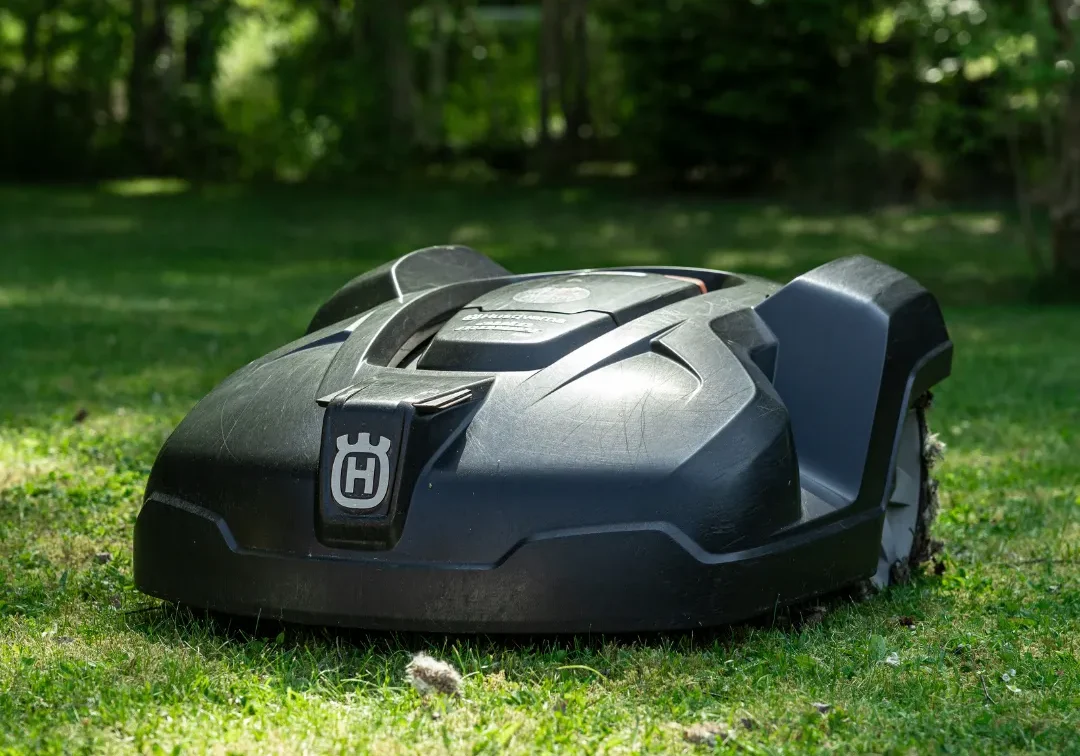 A svart personlig robotgräsklippare med Husqvarna-logo står på grön gräsmatta i en solbelyst trädgård, omgiven av höga träd och frodiga växter i bakgrunden.